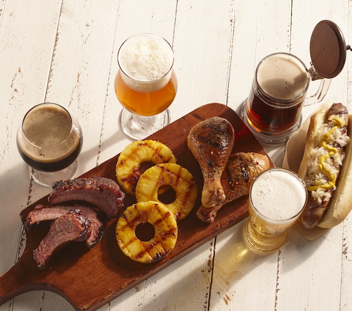 White wooden background with beer, brats, grilled pineapple, chicken, and ribs